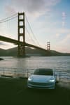 A car with headlights on is parked near the water with the Golden Gate Bridge in the background during sunset