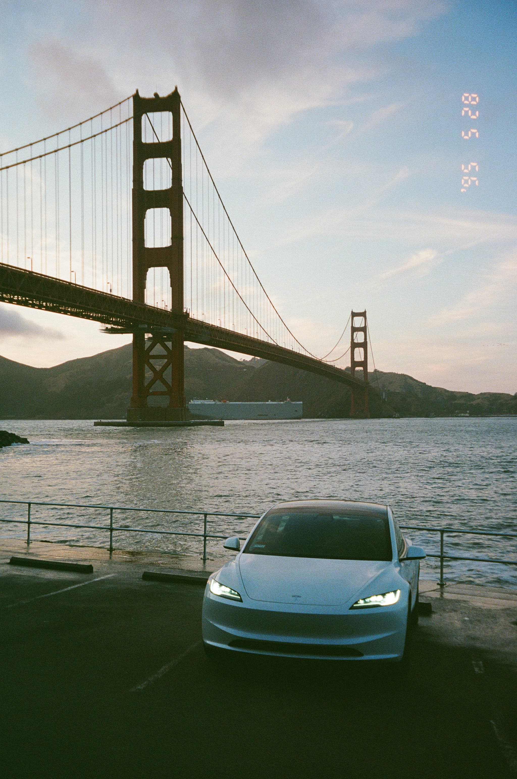 A car with headlights on is parked near the water with the Golden Gate Bridge in the background during sunset