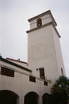 A tall, white bell tower with a red-tiled roof, part of a building with arched windows and a palm tree nearby