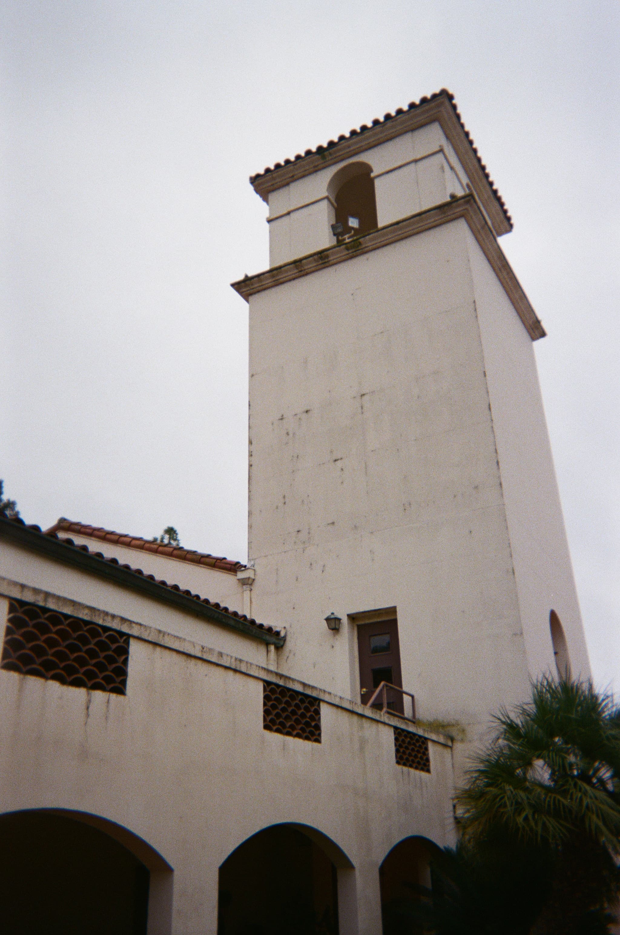 A tall, white bell tower with a red-tiled roof, part of a building with arched windows and a palm tree nearby
