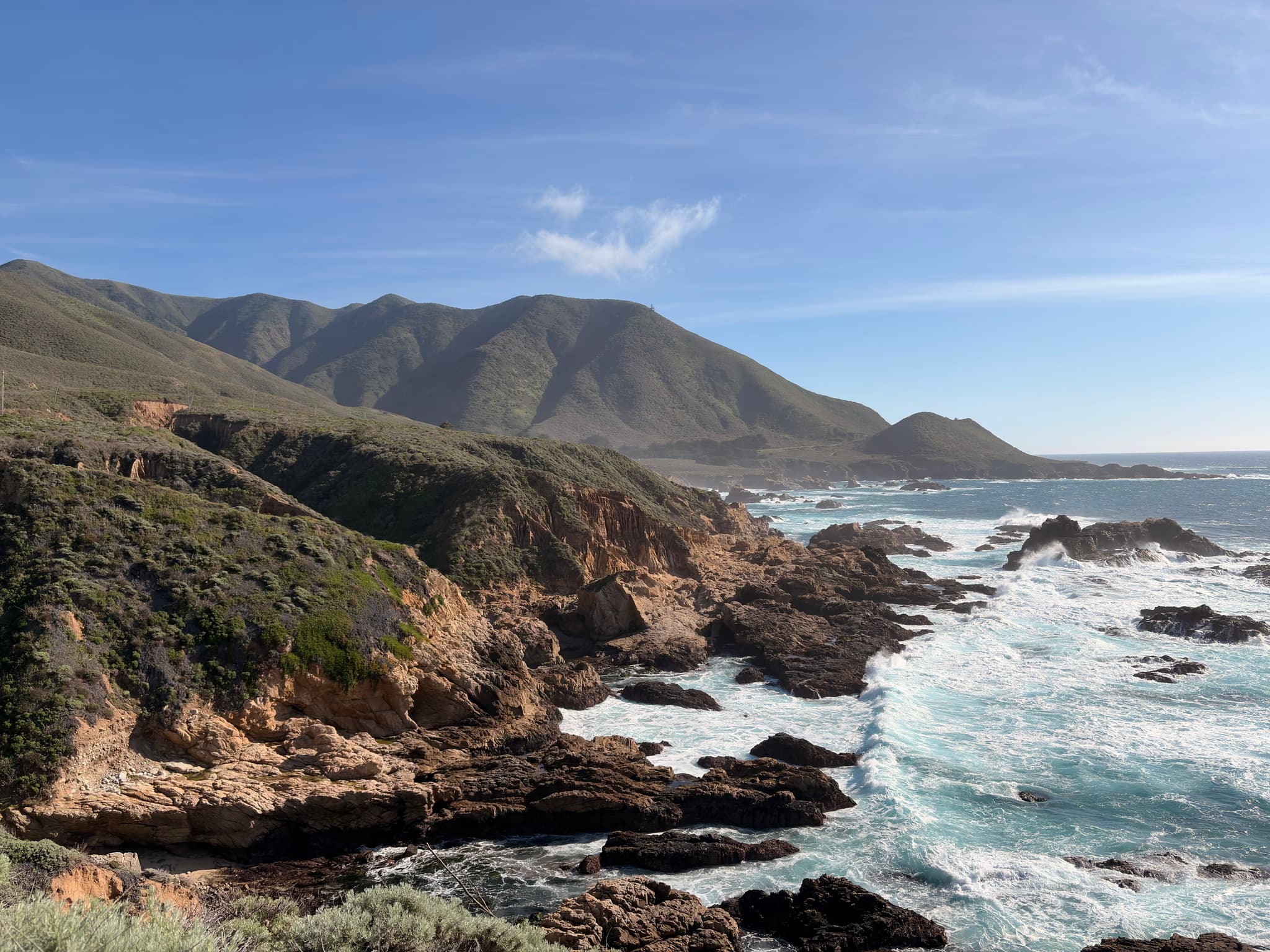 Rocky coastal cliffs and rugged shoreline with waves crashing against rocks, backed by rolling green-brown hills under a clear blue sky
