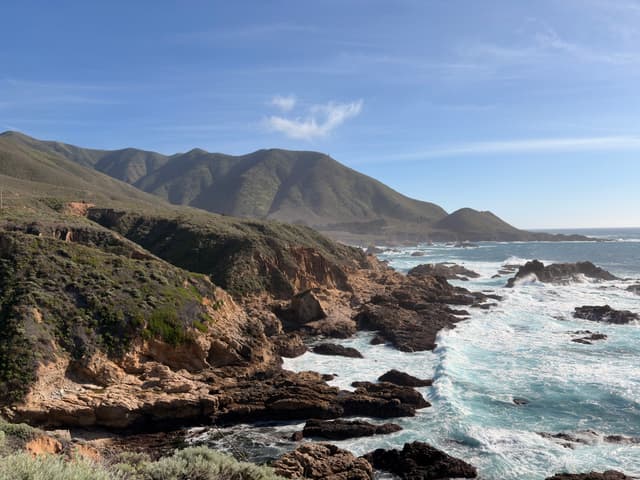 Rocky coastal cliffs and rugged shoreline with waves crashing against rocks, backed by rolling green-brown hills under a clear blue sky