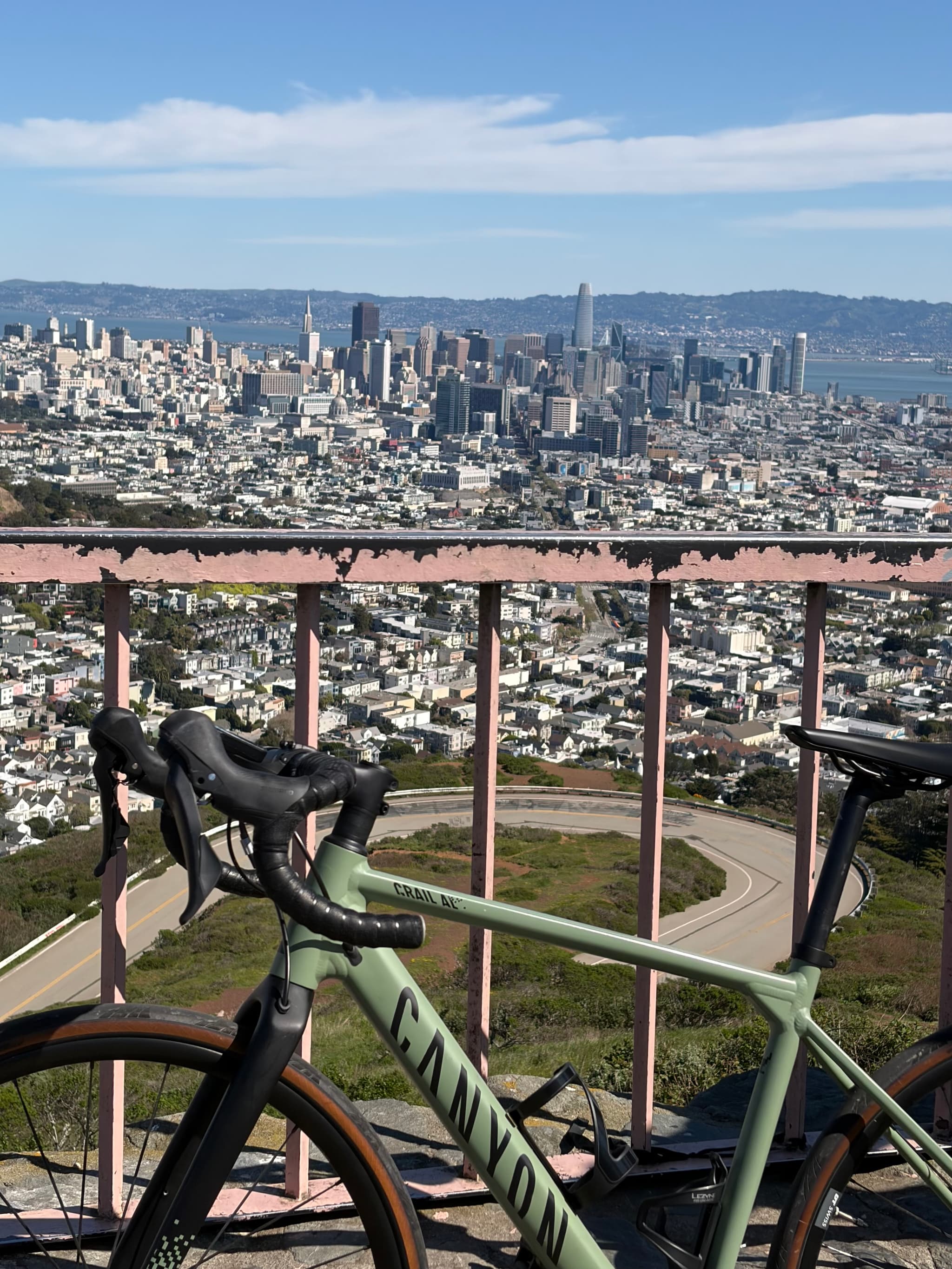 Two bicycles parked at a hilltop overlook with a metal railing, overlooking a dense city skyline and distant hills under a clear blue sky