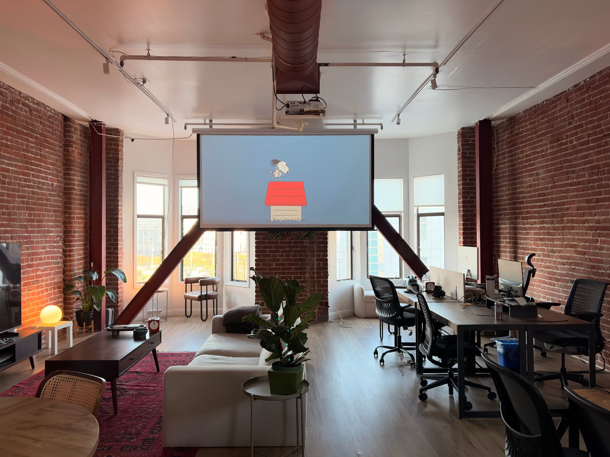 Loft-style coworking office with exposed brick walls, large windows, and desks facing a projector screen showing a login interface