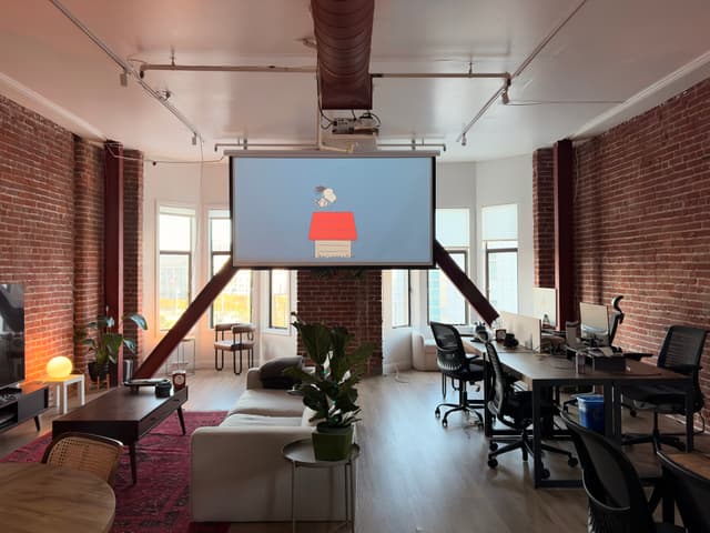 Loft-style coworking office with exposed brick walls, large windows, and desks facing a projector screen showing a login interface