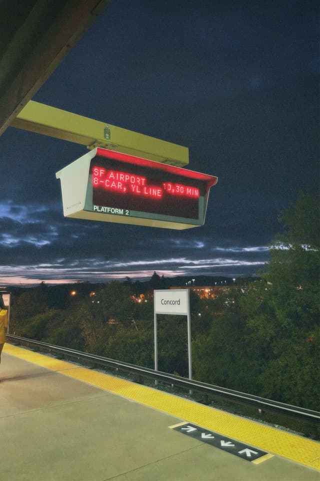 A train platform at night with an electronic sign displaying train information The sky is dark with some clouds, and there are trees and lights in the background