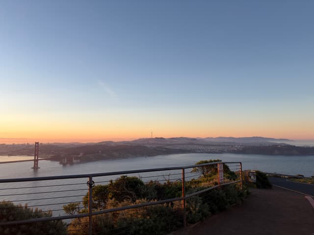 Sunrise view over a calm sea and distant mountains, seen from a coastal lookout with a railing and flowering bushes in the foreground