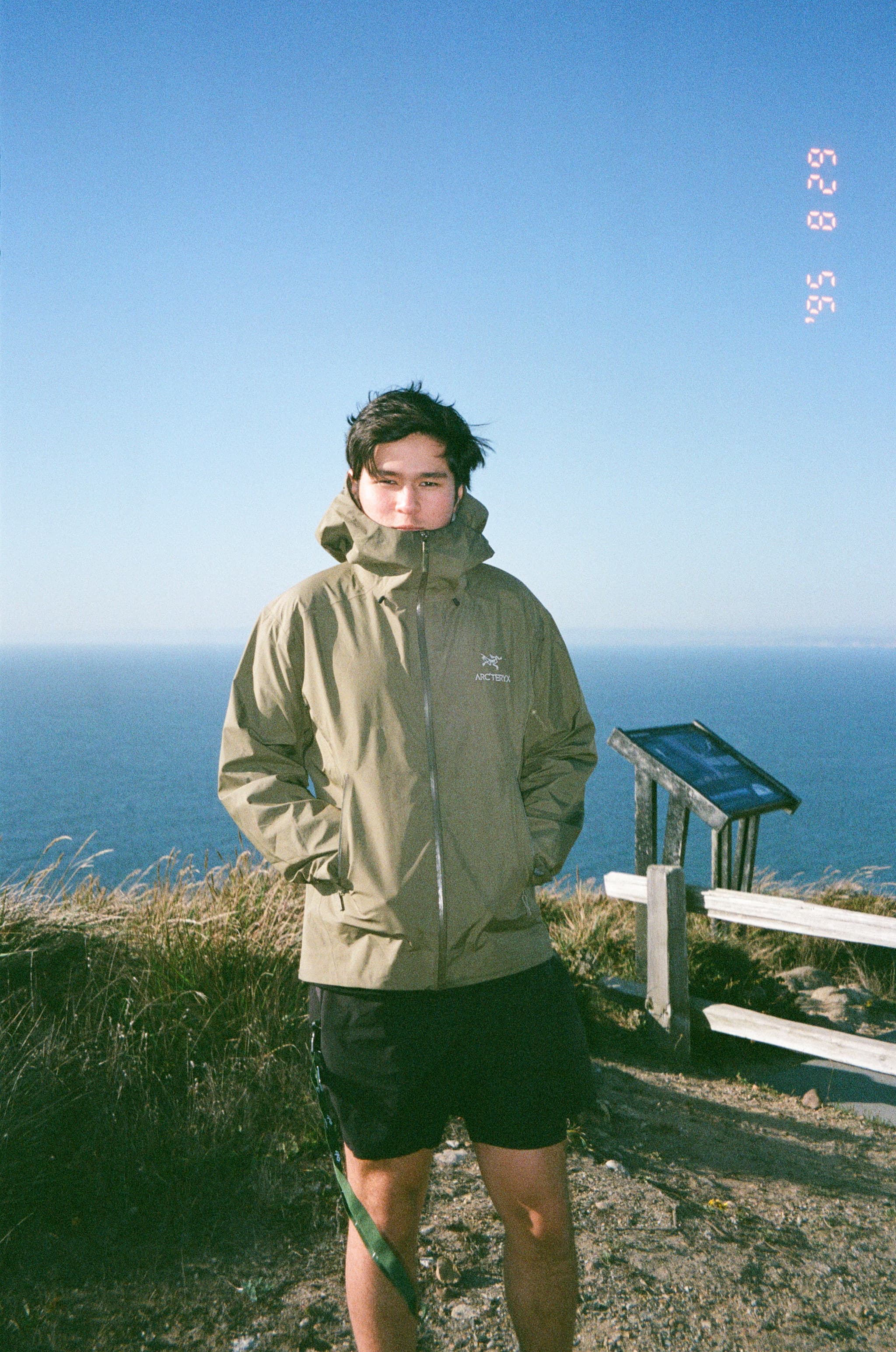 A person wearing a khaki jacket stands near a coastal viewpoint with informational signage, set against a backdrop of the ocean and a clear blue sky