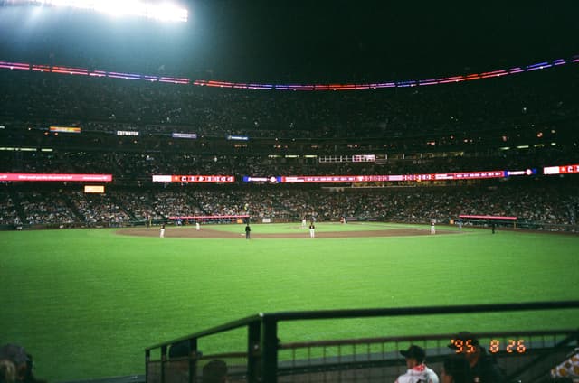 A nighttime baseball game in a stadium with bright lights illuminating the field; players are positioned on the field and the stands are filled with spectators