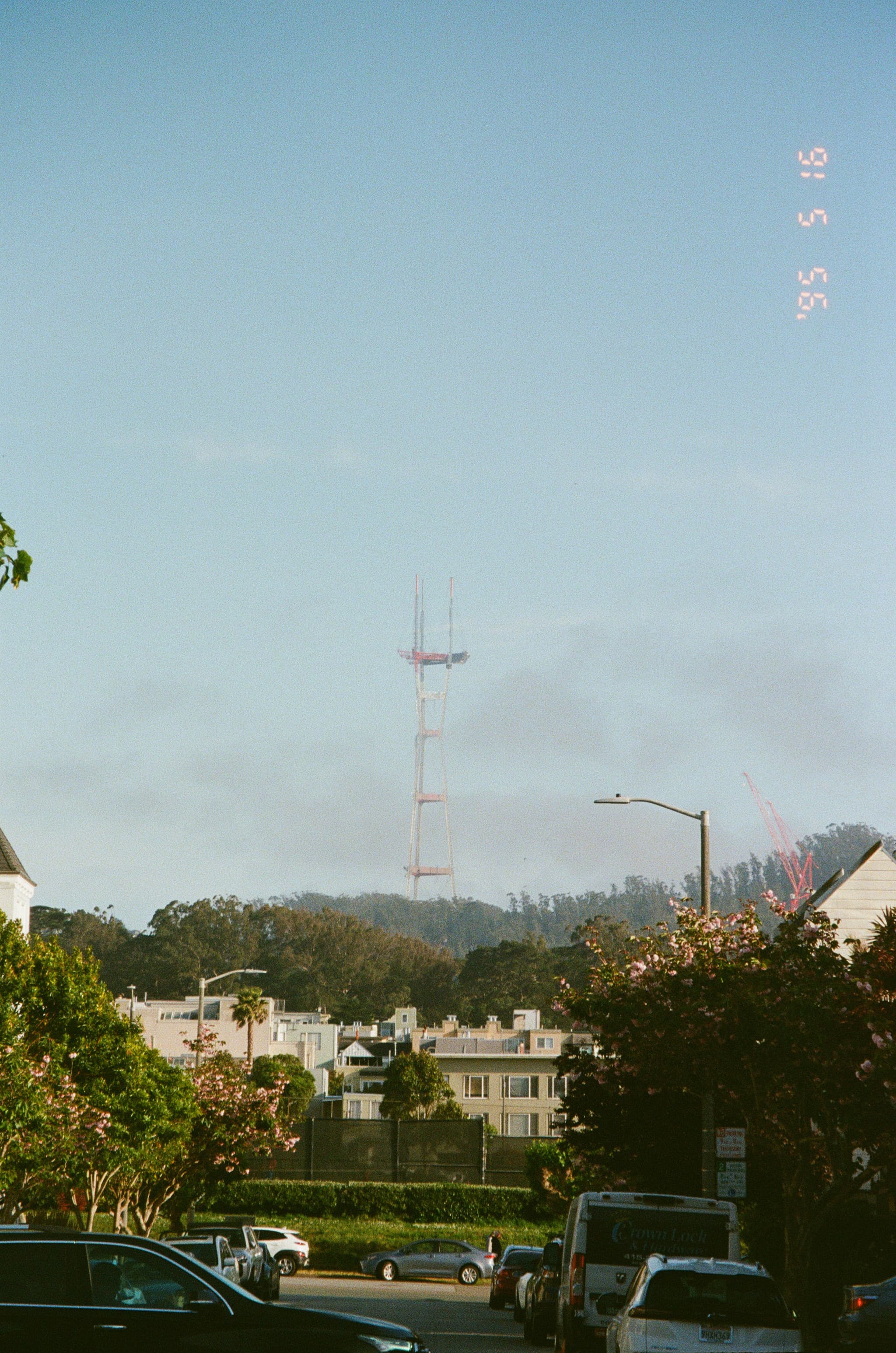 A city street with vehicles and houses in the foreground, trees and hills in the background, and a tall red communication tower rising above the horizon against a clear blue sky