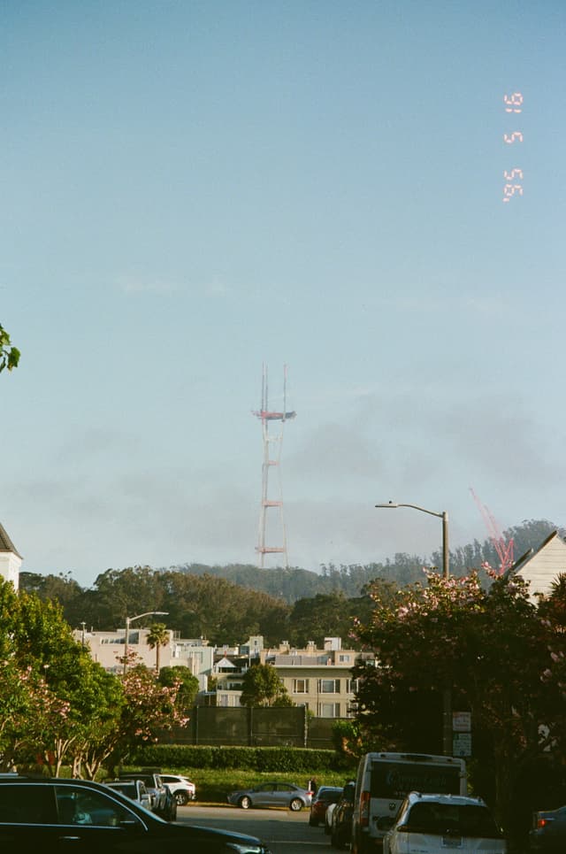 A city street with vehicles and houses in the foreground, trees and hills in the background, and a tall red communication tower rising above the horizon against a clear blue sky