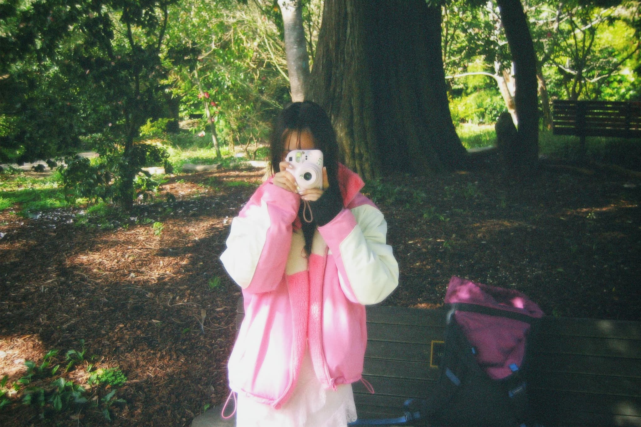 A person in a pink and white jacket is taking a photo with a camera in a park setting, surrounded by trees and greenery A bag is placed on a bench nearby