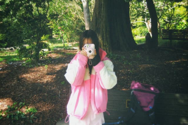 A person in a pink and white jacket is taking a photo with a camera in a park setting, surrounded by trees and greenery A bag is placed on a bench nearby