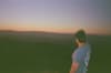 Person in a cap and blue T-shirt looking out over rolling hills at sunset with a hazy horizon