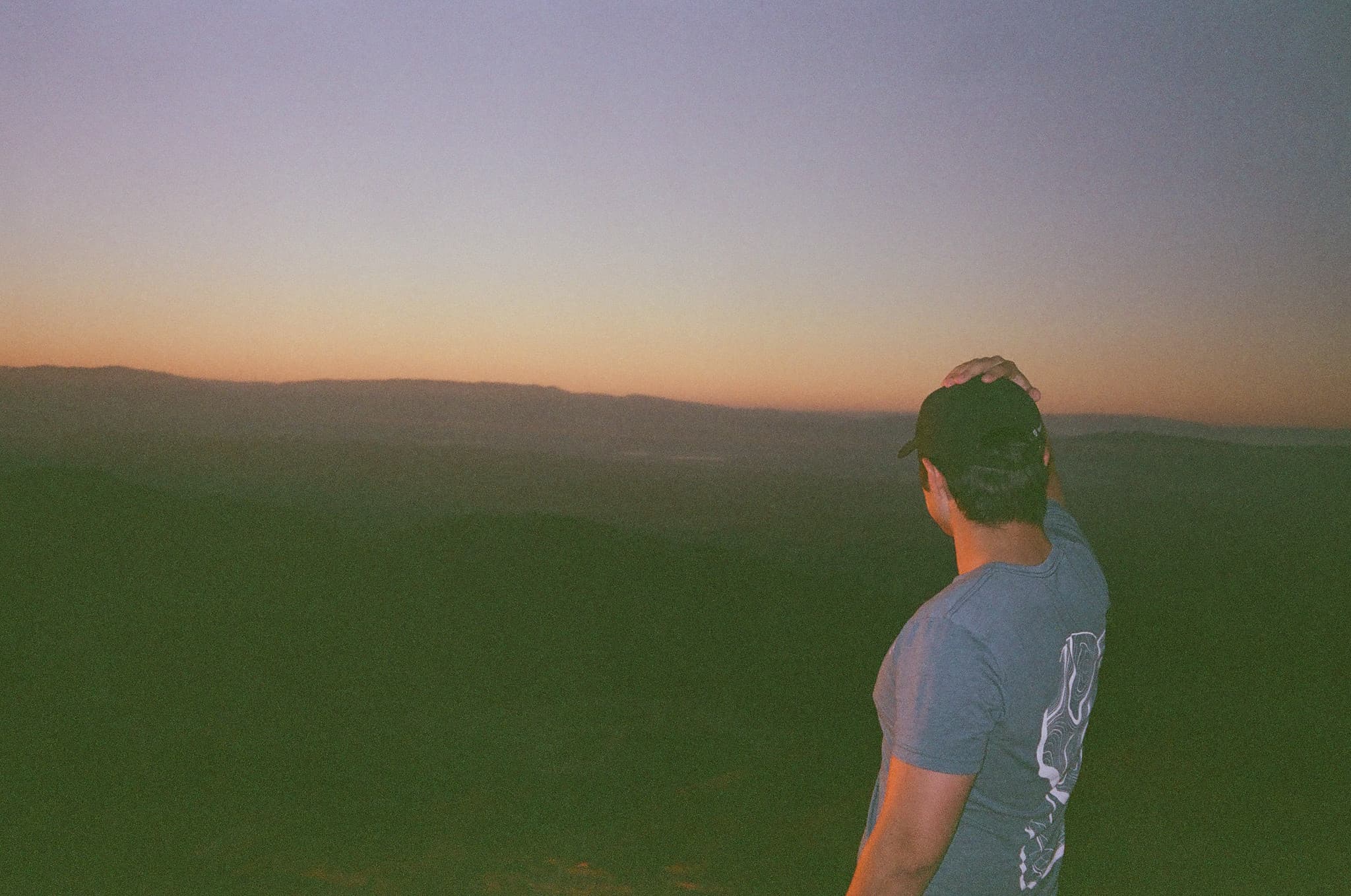 Person in a cap and blue T-shirt looking out over rolling hills at sunset with a hazy horizon