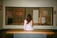 A person with long hair, wearing a light-colored dress, sits on a bench facing a traditional Japanese-style room with sliding doors and tatami mats