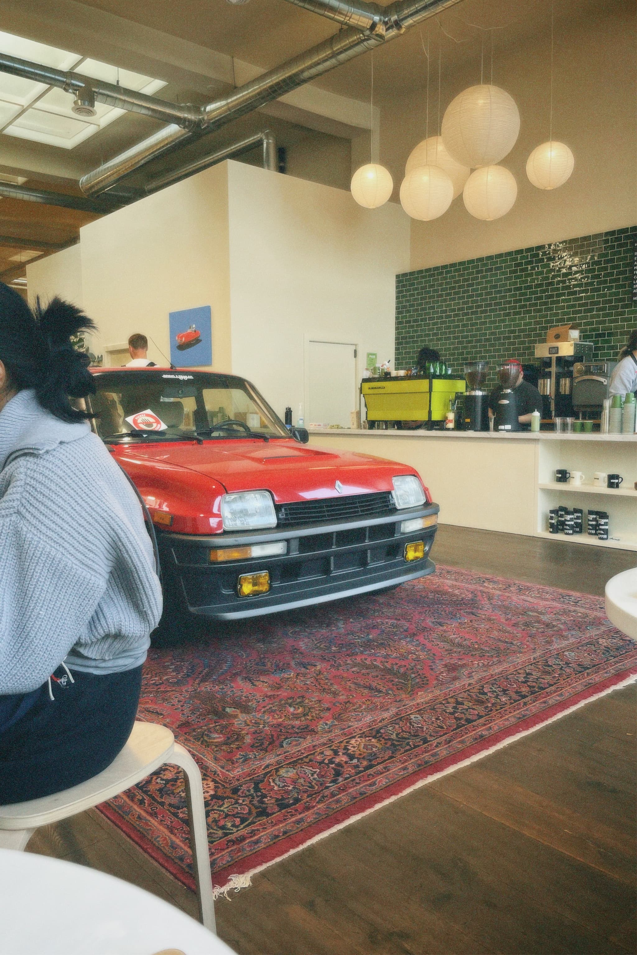 A red car is parked indoors on a decorative rug in a modern cafe setting, with people sitting nearby and a counter in the background