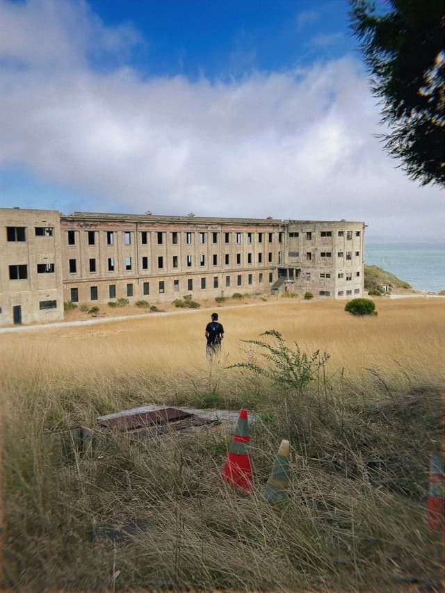 A person stands in a field of tall grass in front of a large, abandoned building near the coast, with traffic cones in the foreground and a cloudy sky above