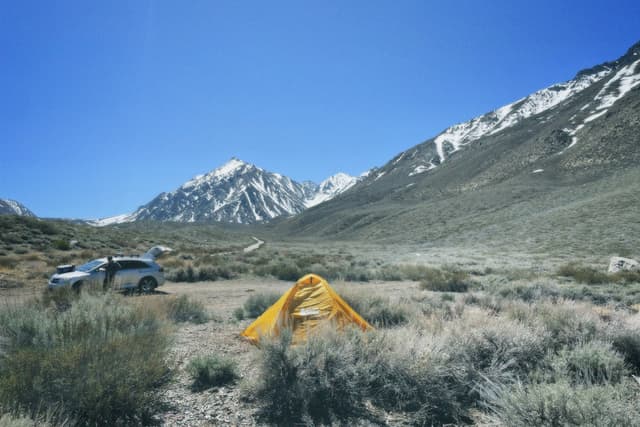A yellow tent set up in a grassy area with a car parked nearby, surrounded by mountains under a clear blue sky