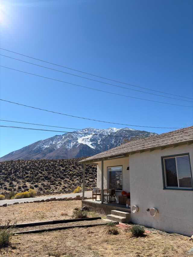 A small house sits in a dry, rocky landscape with a snow-capped mountain in the background under a clear blue sky