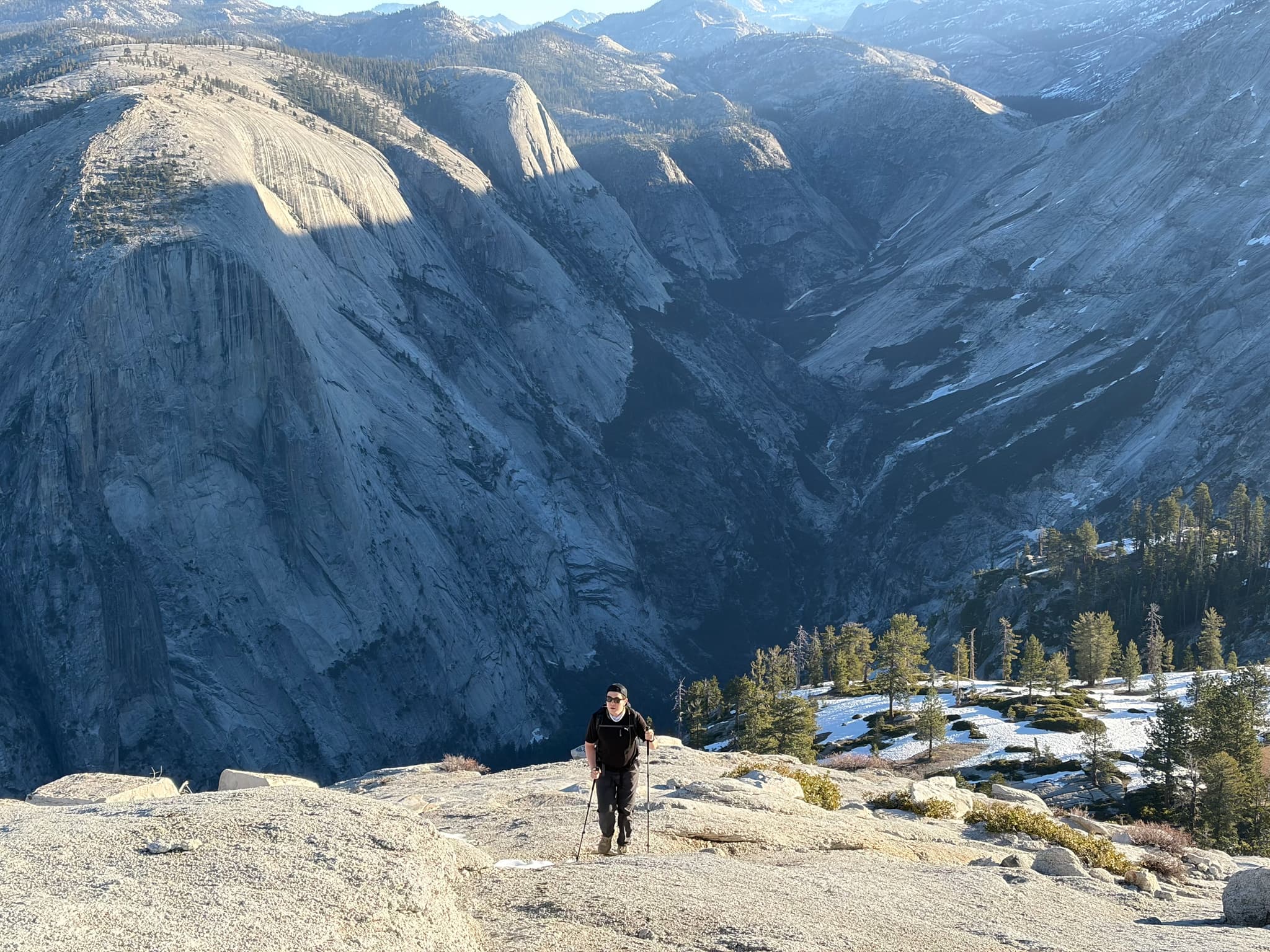 Hiker standing on a granite overlook above a forested valley with distant snow-capped mountains under a clear sky