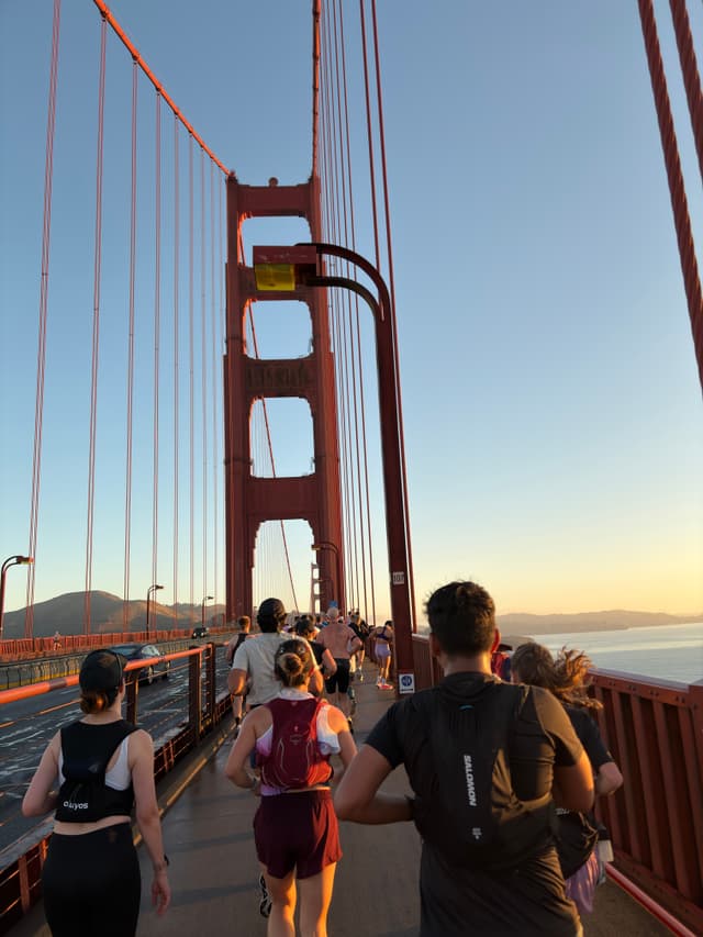 Runners crossing the Golden Gate Bridge at sunrise, with the red suspension towers and cables framing the roadway and bay in the background