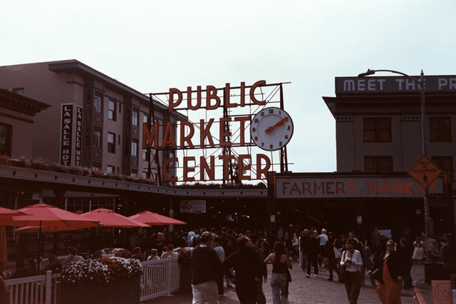 A bustling street scene with a large 'Public Market Center' sign and clock in an urban commercial area, surrounded by buildings and people