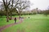 A group of people walking along a path in a lush, green park with large trees and an overcast sky