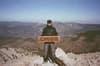 Hiker standing on a rocky mountain summit holding a wooden trail sign, overlooking a vast forested valley and distant ridgelines under a clear blue sky