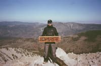 Hiker standing on a rocky mountain summit holding a wooden trail sign, overlooking a vast forested valley and distant ridgelines under a clear blue sky