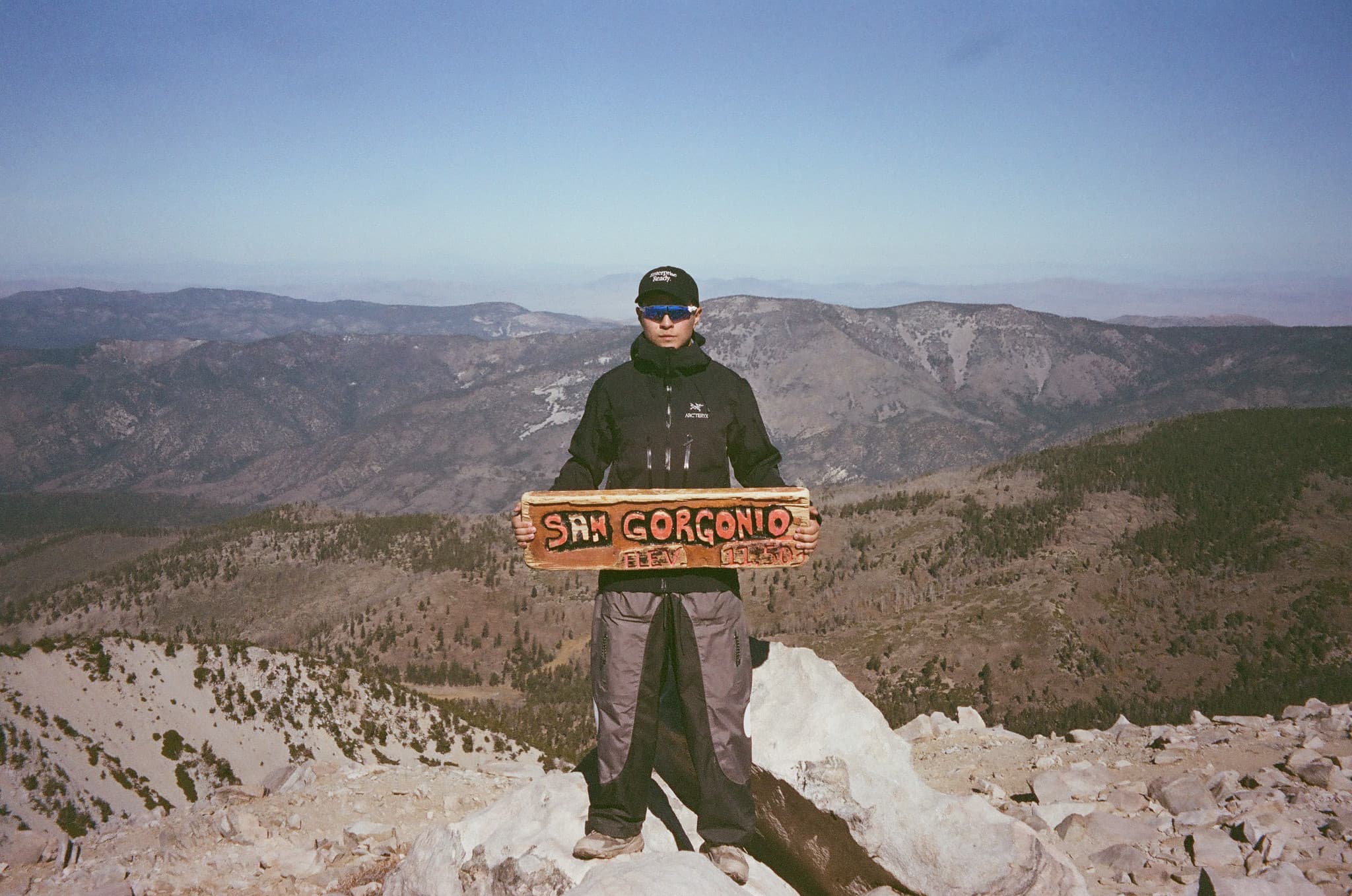 Hiker standing on a rocky mountain summit holding a wooden trail sign, overlooking a vast forested valley and distant ridgelines under a clear blue sky