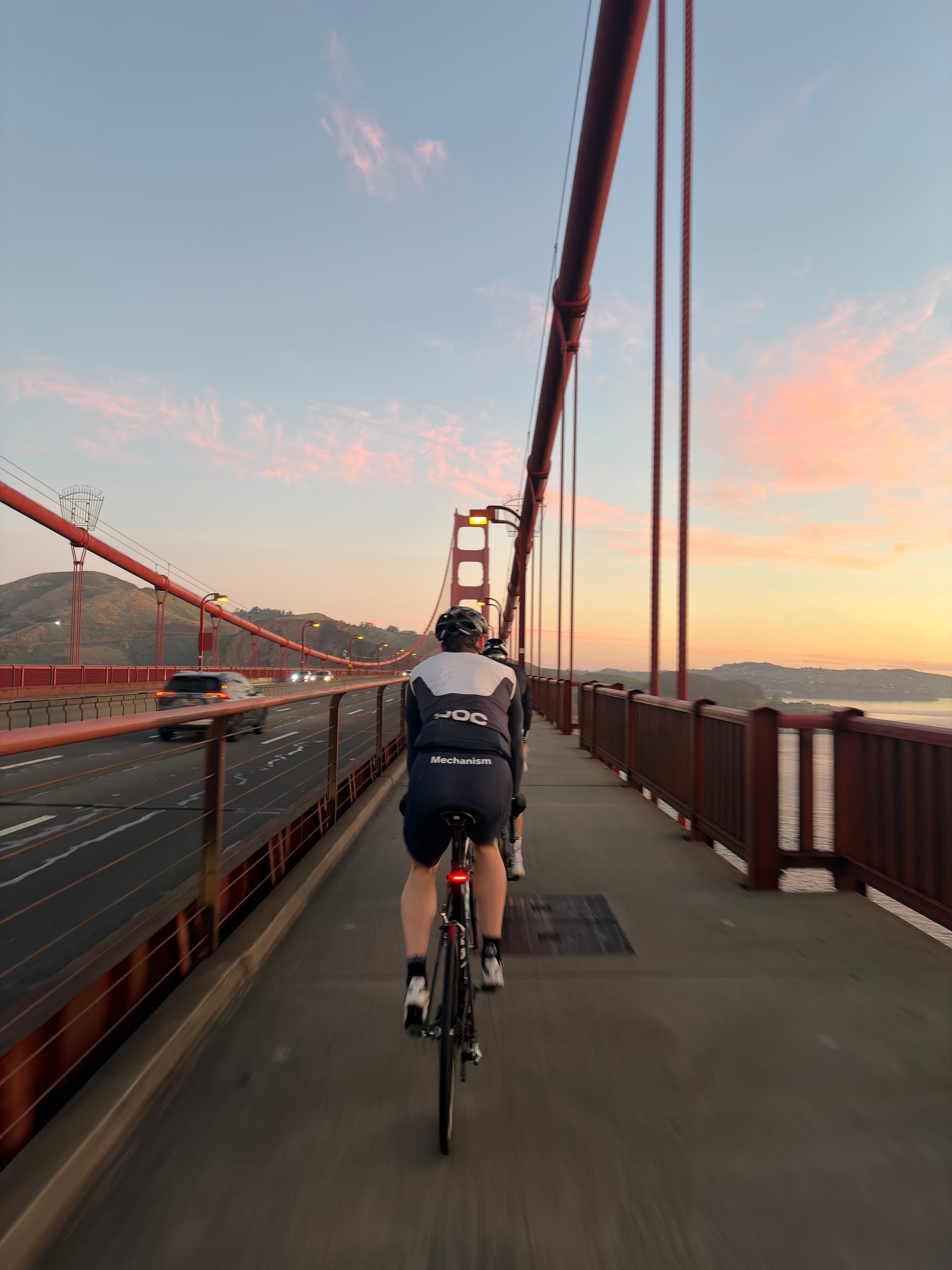 Cyclist riding on the Golden Gate Bridge walkway at sunset, with suspension cables framing the path and distant hills under a pastel sky
