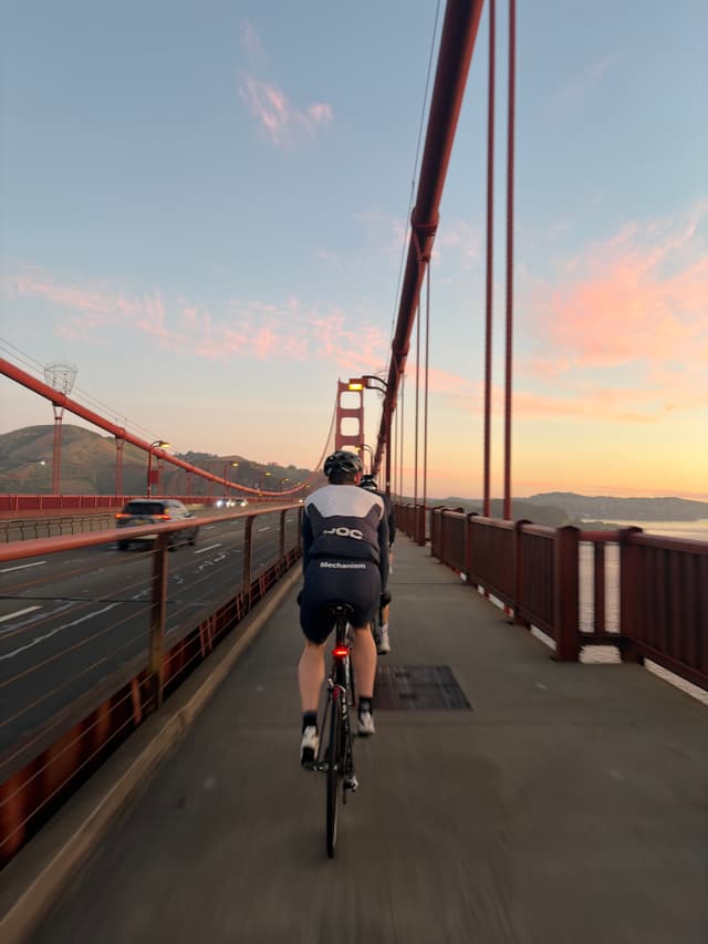 Cyclist riding on the Golden Gate Bridge walkway at sunset, with suspension cables framing the path and distant hills under a pastel sky
