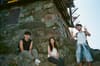 Three people are posing near a stone structure One person is sitting, another is standing with a peace sign, and the third is sitting in the middle The setting appears to be outdoors