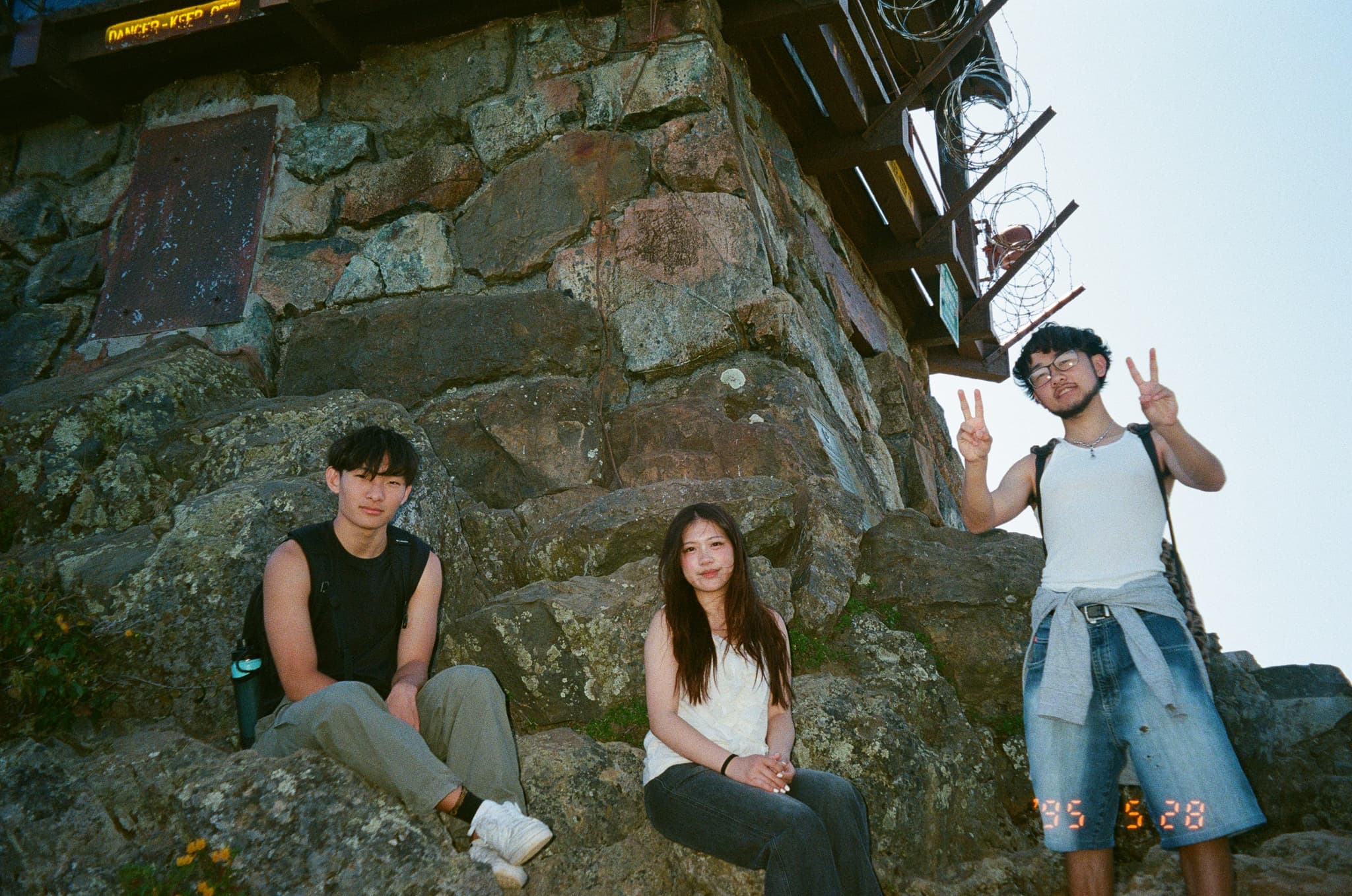 Three people are posing near a stone structure One person is sitting, another is standing with a peace sign, and the third is sitting in the middle The setting appears to be outdoors