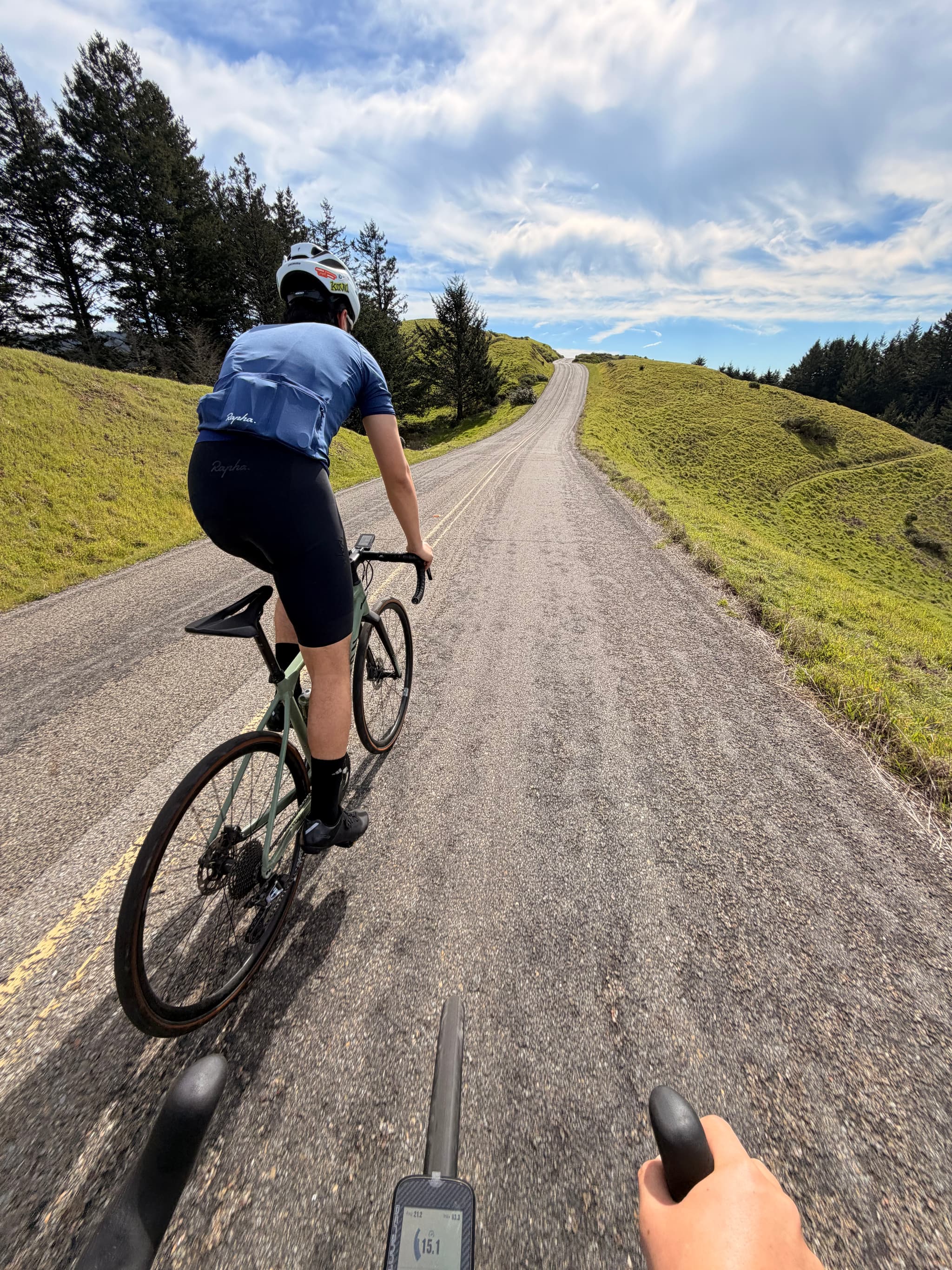 First-person view of a cyclist riding on a gravel country road through rolling green hills under a partly cloudy sky