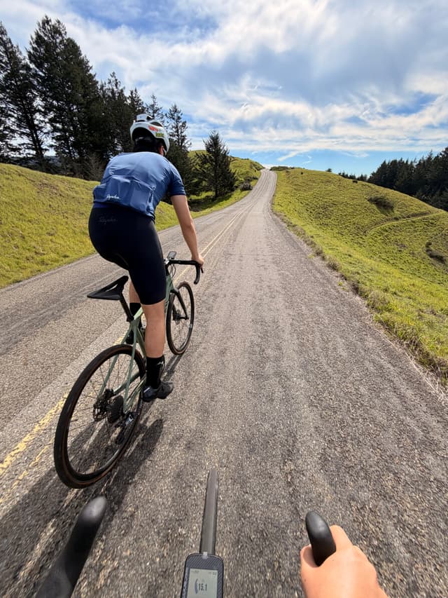 First-person view of a cyclist riding on a gravel country road through rolling green hills under a partly cloudy sky