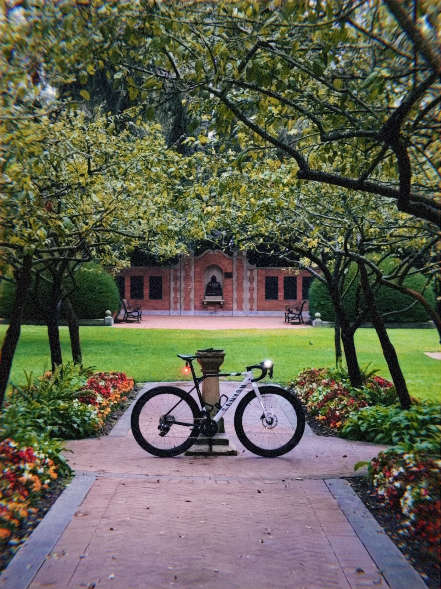 Bicycle parked on a brick walkway framed by flowerbeds and arching trees, with a red brick building in the background
