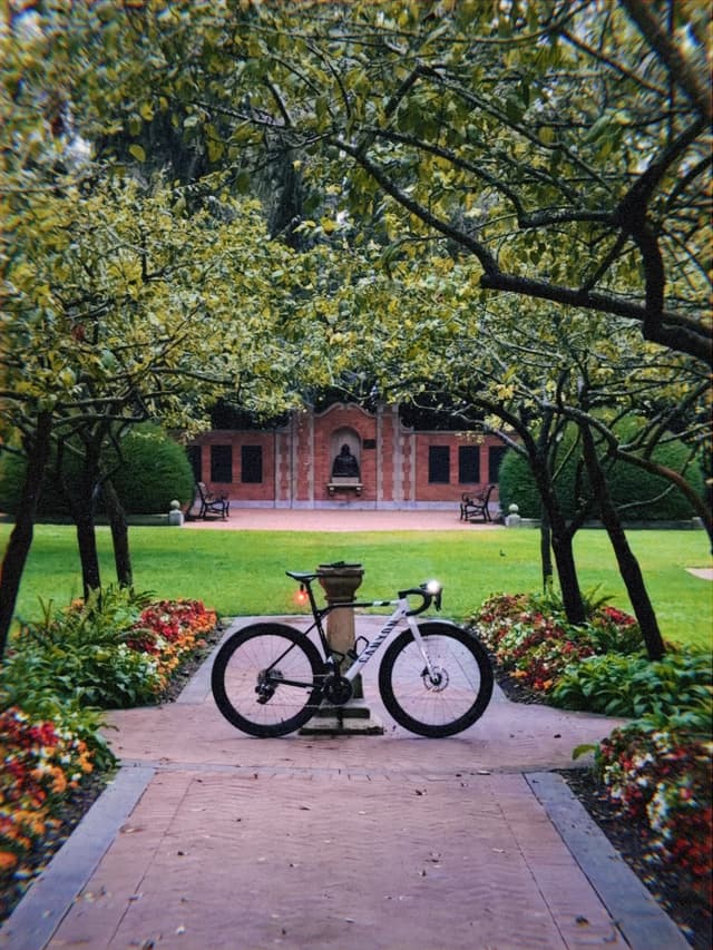 Bicycle parked on a brick walkway framed by flowerbeds and arching trees, with a red brick building in the background