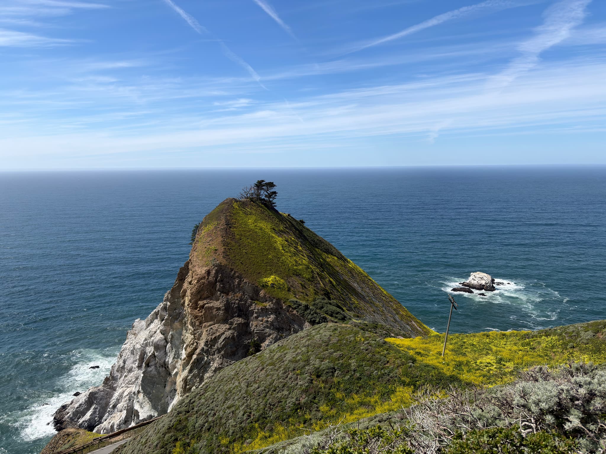 Rocky coastal headland with a steep grassy peak overlooking the ocean under a blue sky with wispy clouds