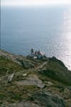A coastal lighthouse perched on a rocky cliff, surrounded by ocean with a pathway leading down from the hillside