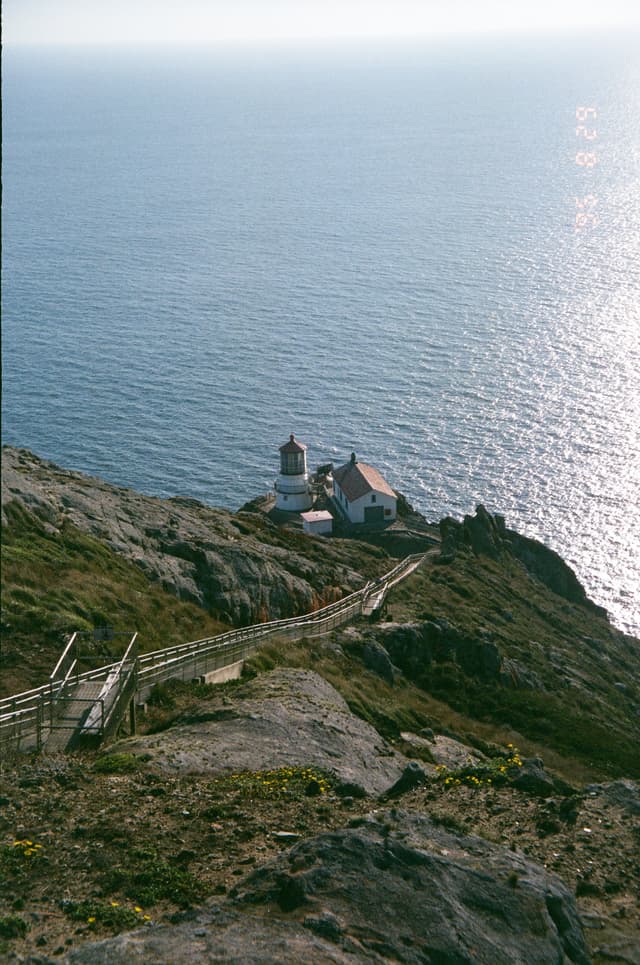 A coastal lighthouse perched on a rocky cliff, surrounded by ocean with a pathway leading down from the hillside