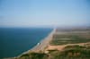 A vast coastal landscape with a long stretch of sandy beach bordered by the sea on one side and green vegetation on the other, under a clear blue sky