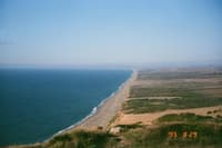 A vast coastal landscape with a long stretch of sandy beach bordered by the sea on one side and green vegetation on the other, under a clear blue sky