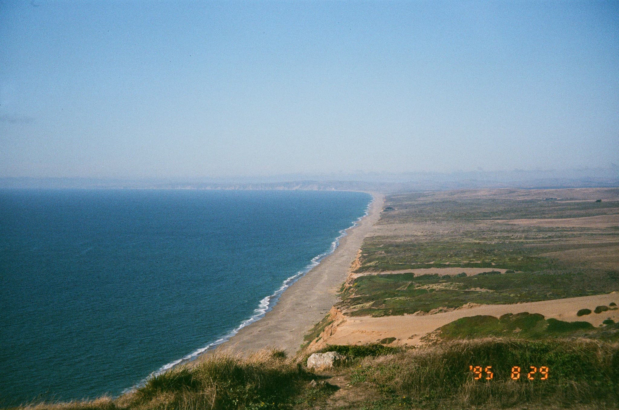 A vast coastal landscape with a long stretch of sandy beach bordered by the sea on one side and green vegetation on the other, under a clear blue sky