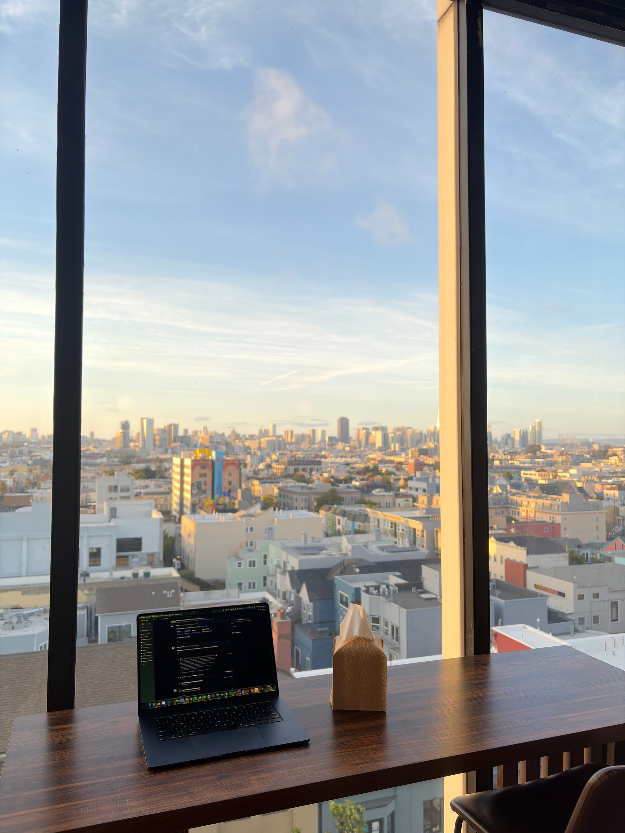 Laptop and takeaway coffee cup on a wooden desk by a large window overlooking a sunlit city skyline and river
