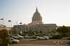 A large neoclassical building with a prominent dome, surrounded by flags and trees, with cars parked in the foreground