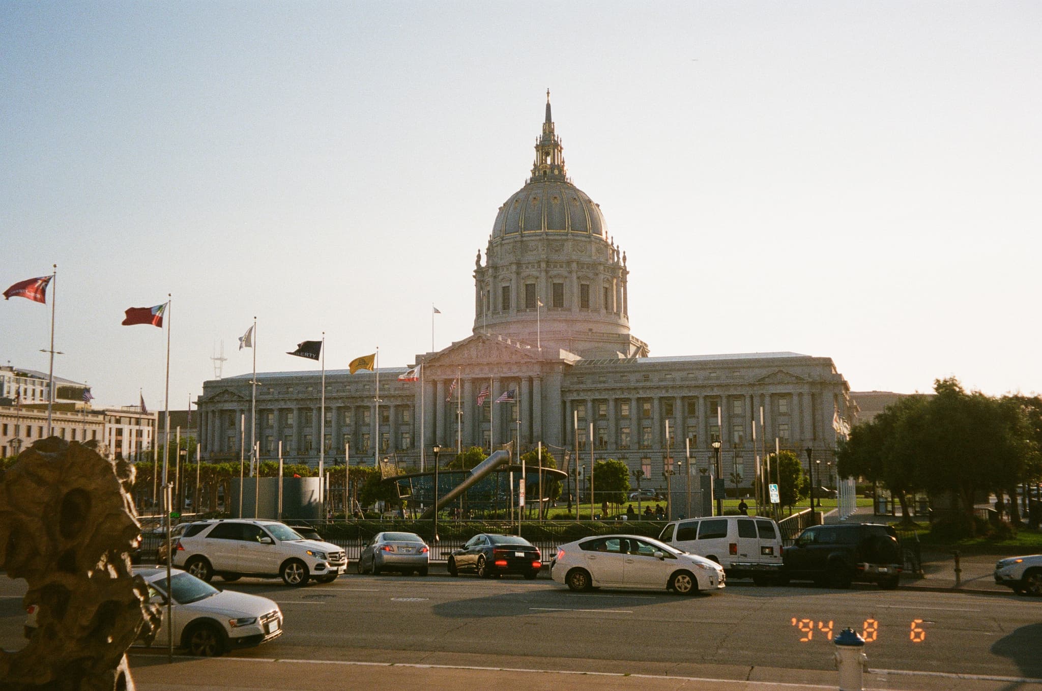 A large neoclassical building with a prominent dome, surrounded by flags and trees, with cars parked in the foreground