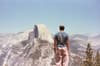A person stands on a rocky ledge overlooking a vast mountain landscape with a distinctive granite dome in the distance under a clear blue sky