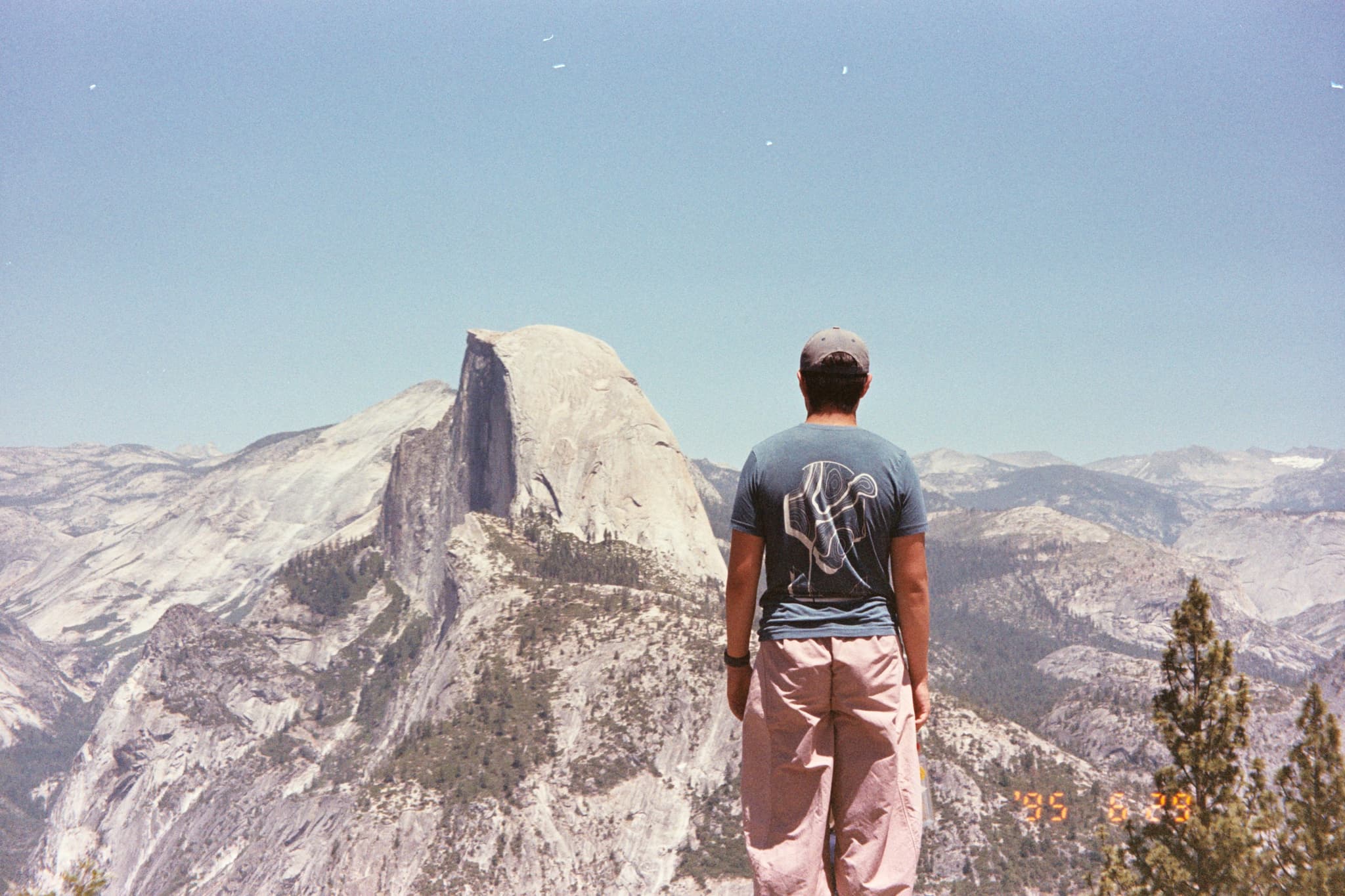 A person stands on a rocky ledge overlooking a vast mountain landscape with a distinctive granite dome in the distance under a clear blue sky